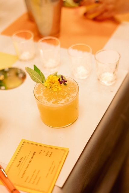 A finished golden cocktail garnished with an edible flower beside its Shaken build card